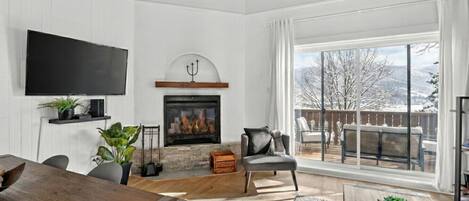 A living room with a fireplace, a TV mounted on the wall, and a large window overlooking a deck.