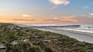 Een strand met een houten pier die zich uitstrekt over de oceaan, omringd door grasduinen en enkele gebouwen in de verte.