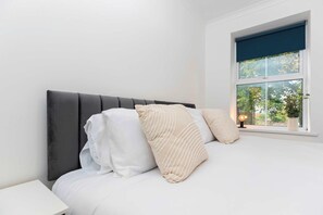 A bedroom with a bed, white bedding, a dark headboard, and a window with a view of trees.