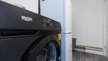 A front view of a washing machine with a control panel, a white refrigerator, and a trash bin in a laundry room.