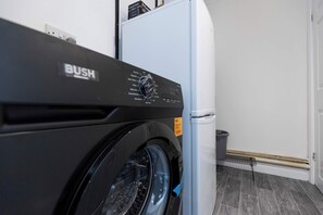 A front view of a washing machine with a control panel, a white refrigerator, and a trash bin in a laundry room.