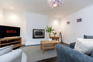A modern living room with a flat-screen TV displaying 'NETFLIX', a wooden coffee table, a potted plant, a floor lamp, and a blue sofa.