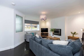A modern living room with a grey sofa, a flat-screen TV, and a decorative chandelier. - Central Bishop - 4 Beds, Games Room, Secure Parking (Bishop Auckland)