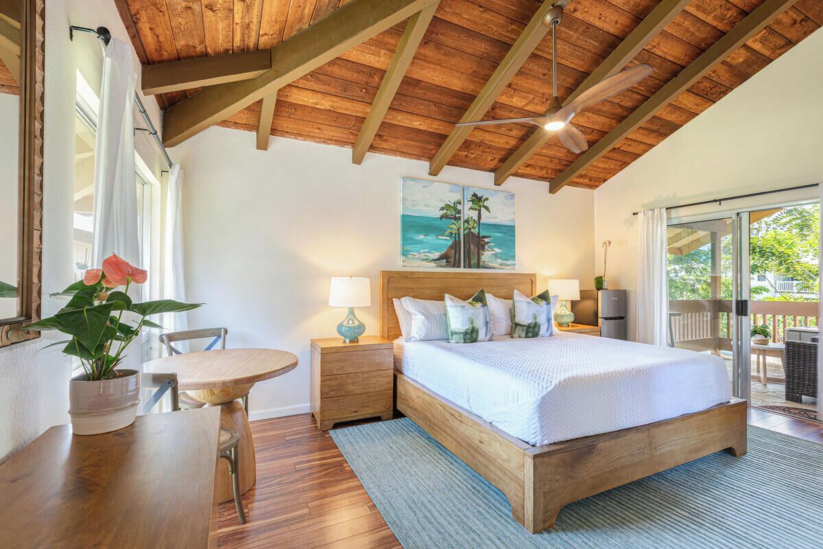 A bedroom with a wooden ceiling, a bed with white bedding, a painting of a beach scene, and a view of the outdoors through a sliding door.