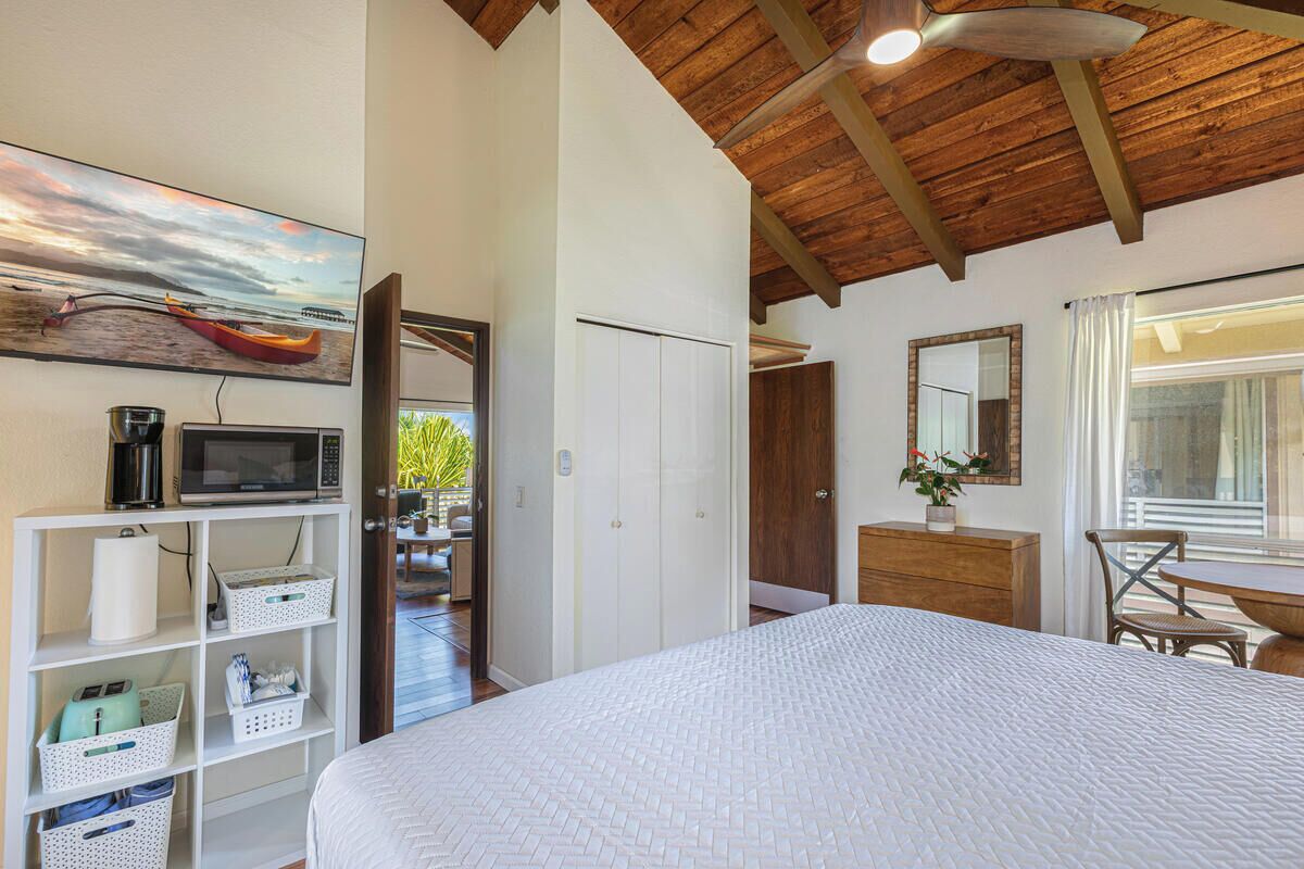 Une chambre avec un lit, un plafond en bois, une étagère avec des paniers de rangement, un four à micro-ondes, un tableau, une porte donnant sur un balcon et une fenêtre avec des rideaux.