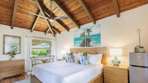 Una camera da letto con soffitto in legno, un letto con biancheria bianca, un comodino con una lampada, un frigorifero e un quadro raffigurante una spiaggia.