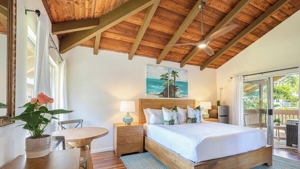 Una camera da letto con soffitto in legno, un letto con biancheria bianca, un quadro raffigurante una spiaggia e vista all'esterno tramite una porta scorrevole.
