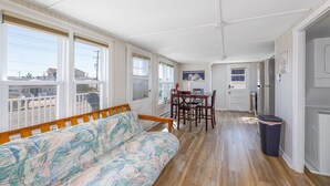 A room with a wooden floor, a sofa, a dining table with chairs, and a trash bin.