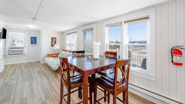 A spacious room with a wooden dining table and chairs, a bed with a floral bedspread, and large windows offering a view of the city.