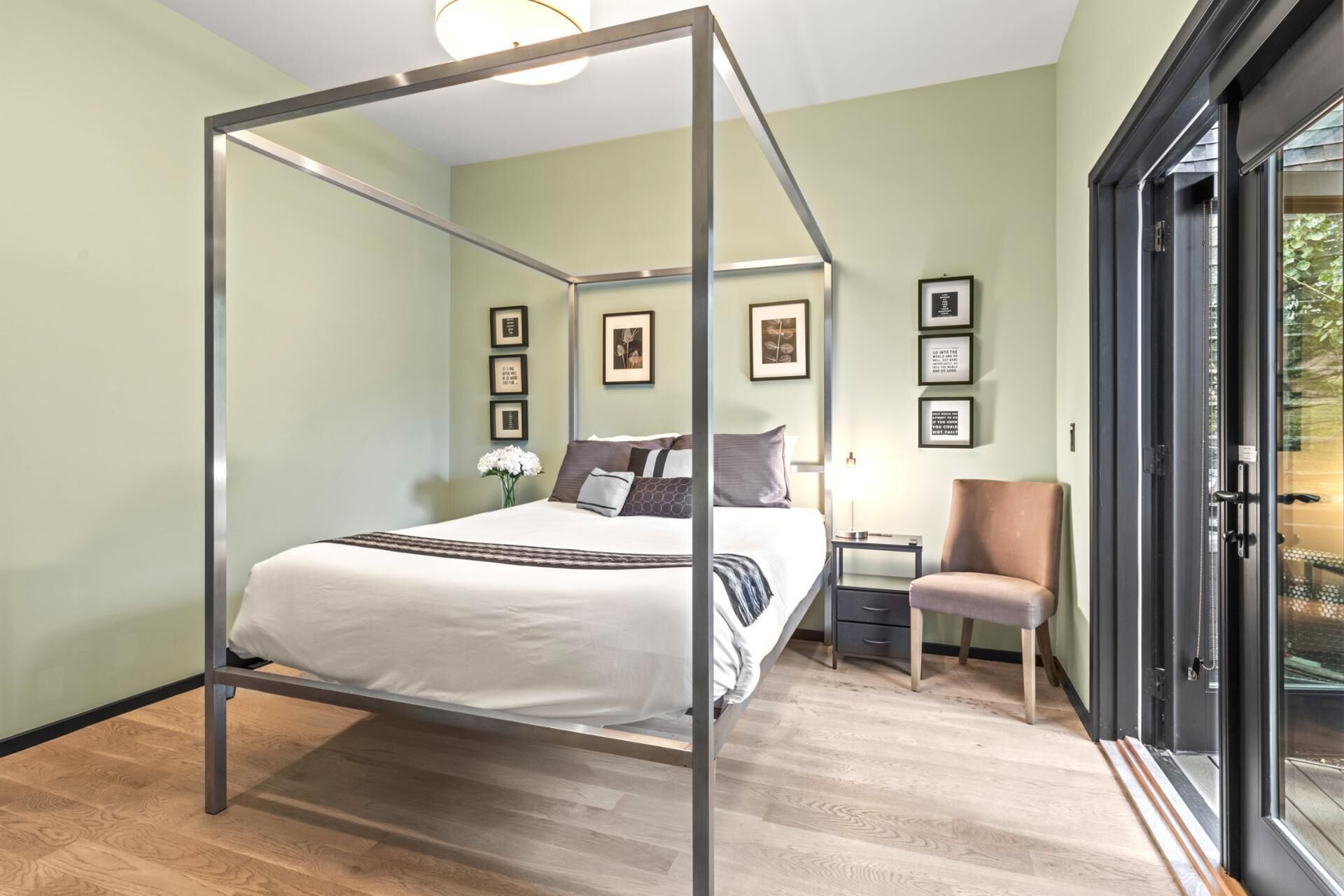 A four-poster bed in a room with light green walls, framed pictures, a small table with a lamp, and a chair.