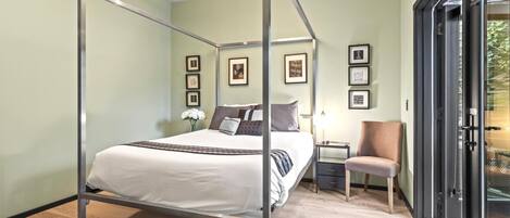 A four-poster bed in a room with light green walls, framed pictures, a small table with a lamp, and a chair.