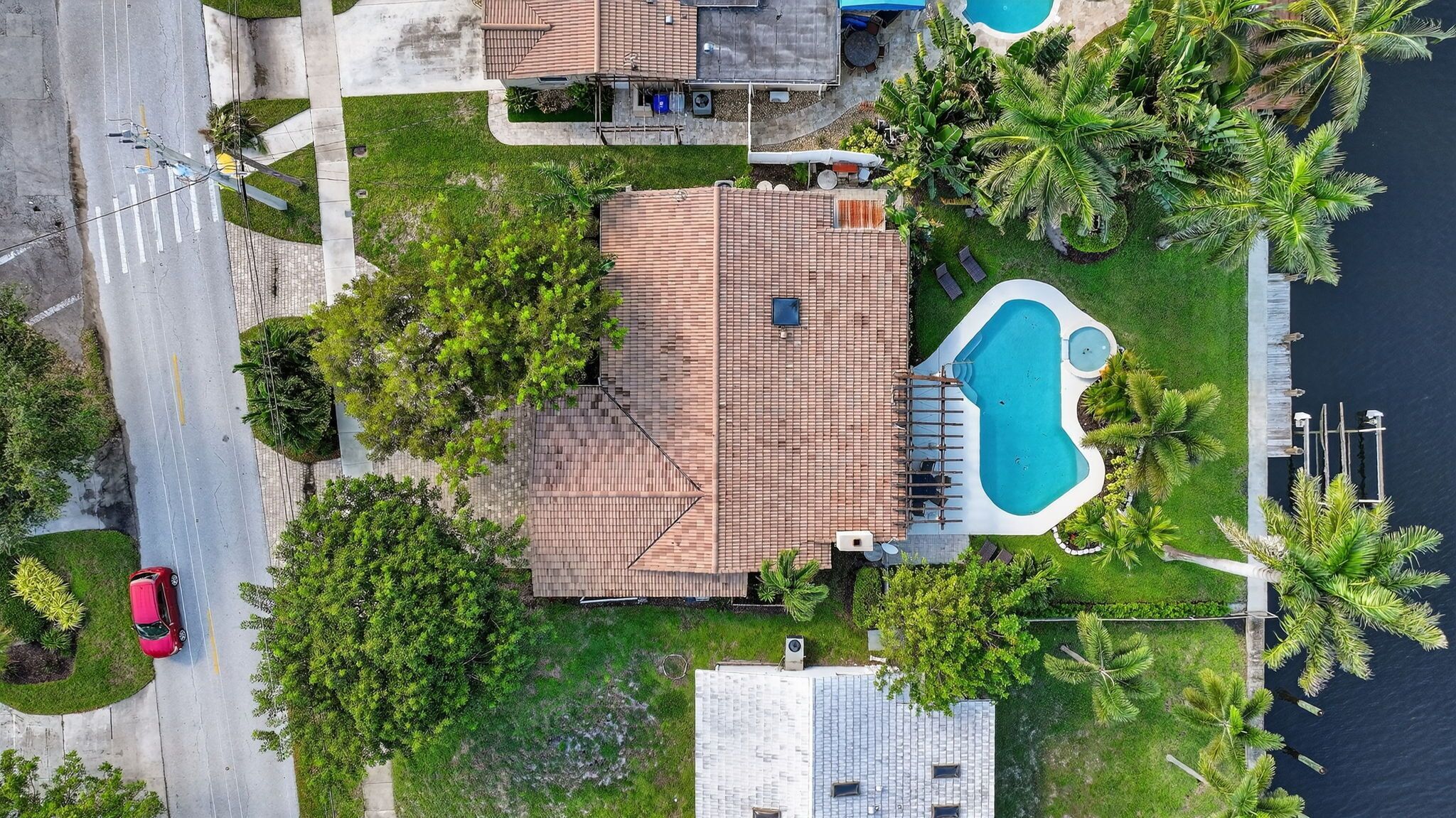 An aerial view of a residential area with a swimming pool, a red car parked on the street, and lush greenery.