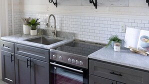 A kitchen with dark cabinets, a stainless steel sink, and a built-in oven. There are shelves above the sink holding various kitchen items and decorations. - Charming Apt. with Marble Mantel (Troy)