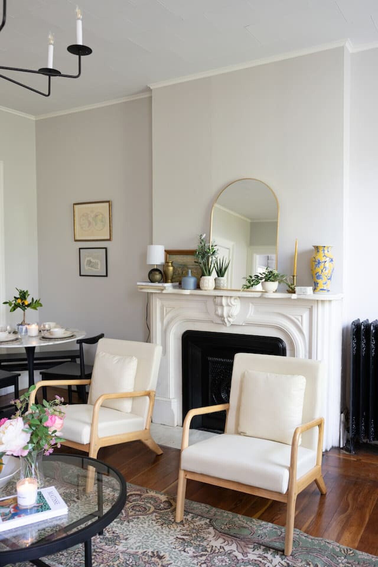 Uma sala de estar com lareira, dois poltronas brancas, uma mesa de centro redonda com um vaso de flores e um espaço de refeições com uma mesa posta para dois.