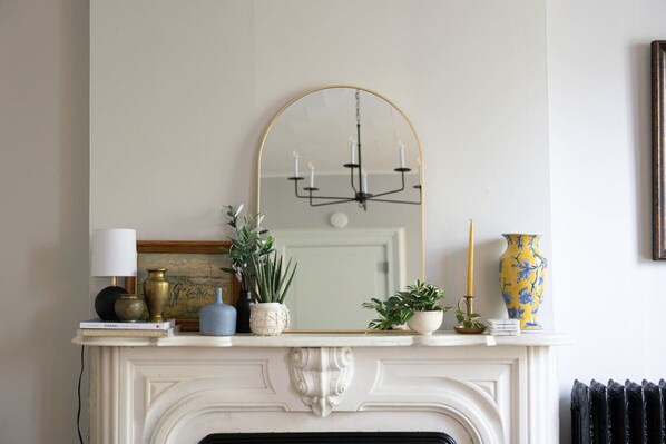 A white fireplace mantel with a mirror above it, a chandelier, and various decorative items including a vase, a candle, and a book.