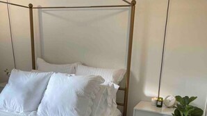 A four-poster bed with white bedding, a bedside table with a lamp, and a potted plant.