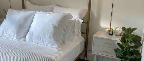 A four-poster bed with white bedding, a bedside table with a lamp, and a potted plant.