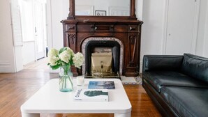 Ein Wohnzimmer mit Kamin, einer schwarzen Ledersofa, einem weißen Couchtisch mit Blumenvase und Zeitschrift, und einer weißen Tür im Hintergrund.