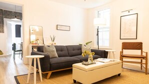 A modern living room with a grey sofa, a white ottoman, and a wooden chair. There are two small side tables, a floor lamp, and a framed picture on the wall. - Peaceful, Artsy North Troy Riverview Apt (Troy)
