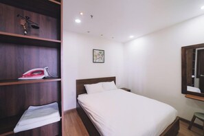 A bedroom with a bed, a wooden shelf with an ironing board and clothes, a framed picture on the wall, and a mirror on the wall. - Dalat Center Residence (Dalat)