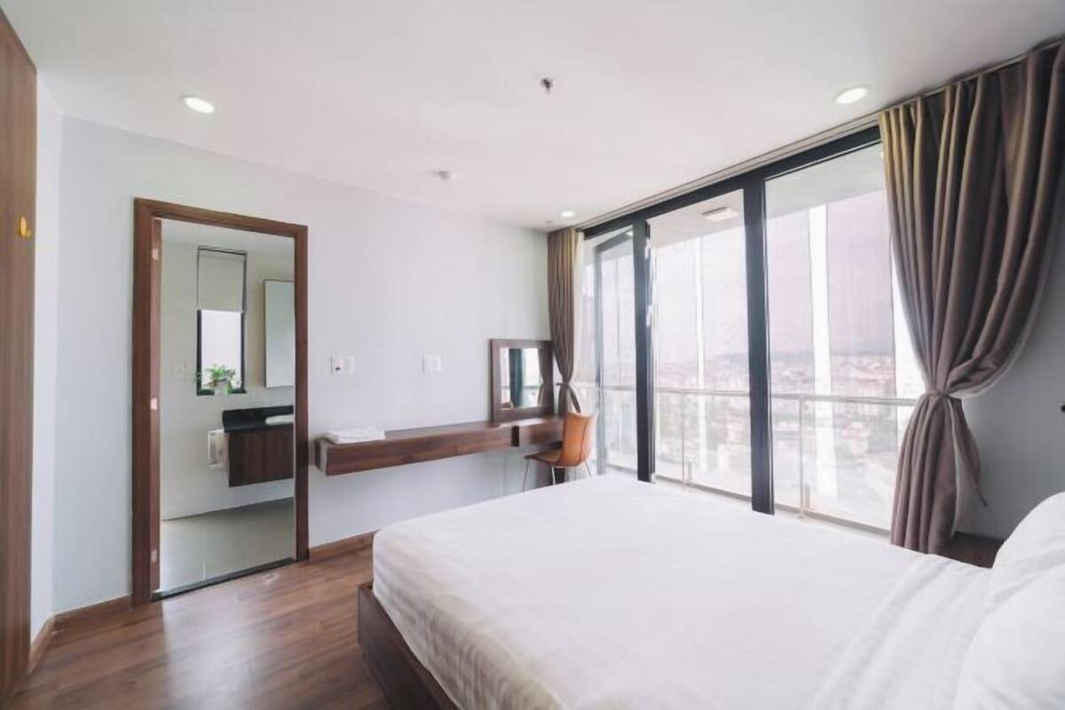 A modern hotel room with a large bed, a wooden floor, a bathroom with a mirror, and a large window with curtains.