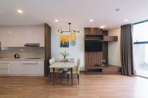 A modern kitchen with white cabinets, a dining table with chairs, a pendant light, and a flat-screen TV mounted on a wooden panel. - Dalat Center Residence (Dalat)