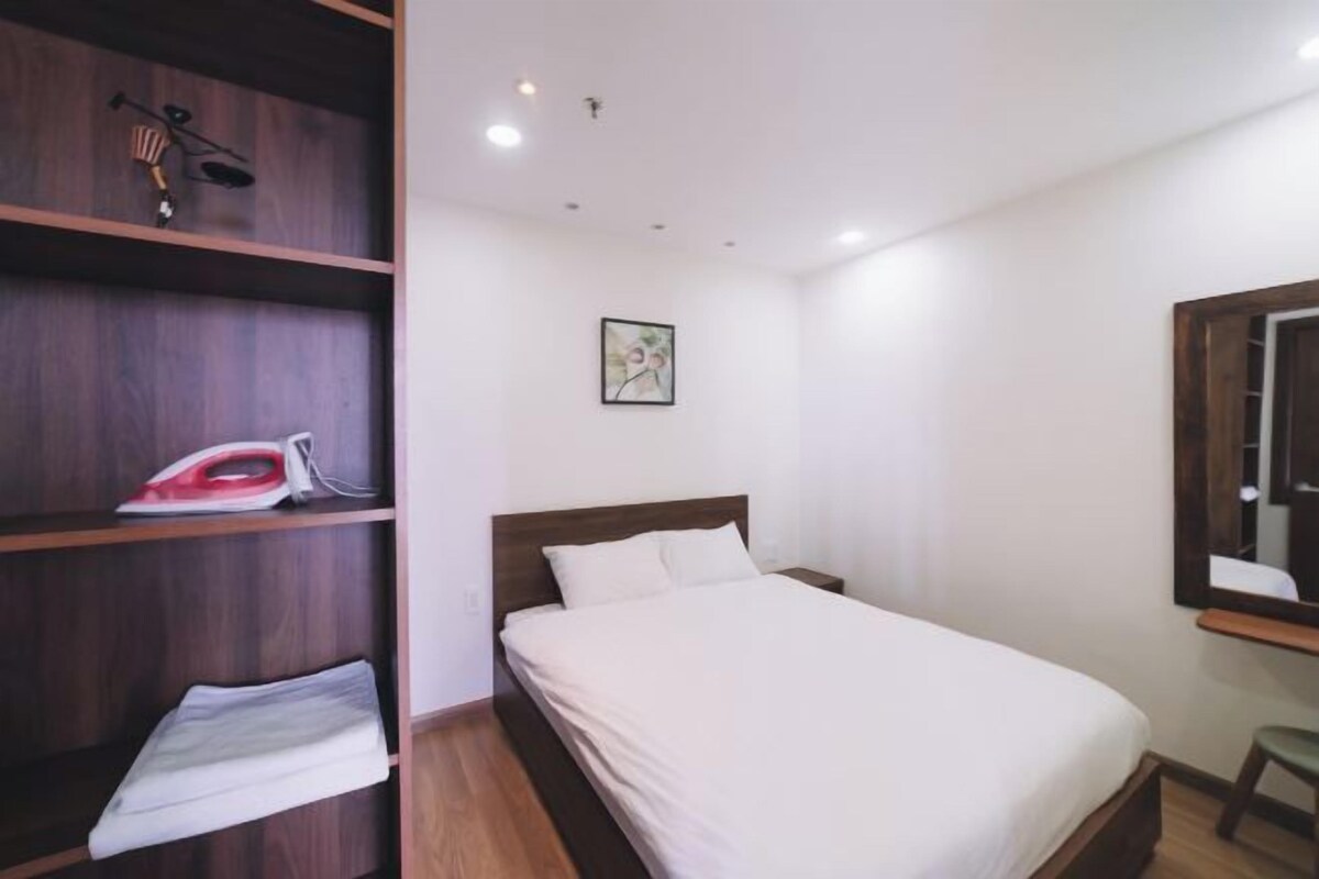A bedroom with a wooden bed, a shelf with an ironing board, and a framed picture on the wall.