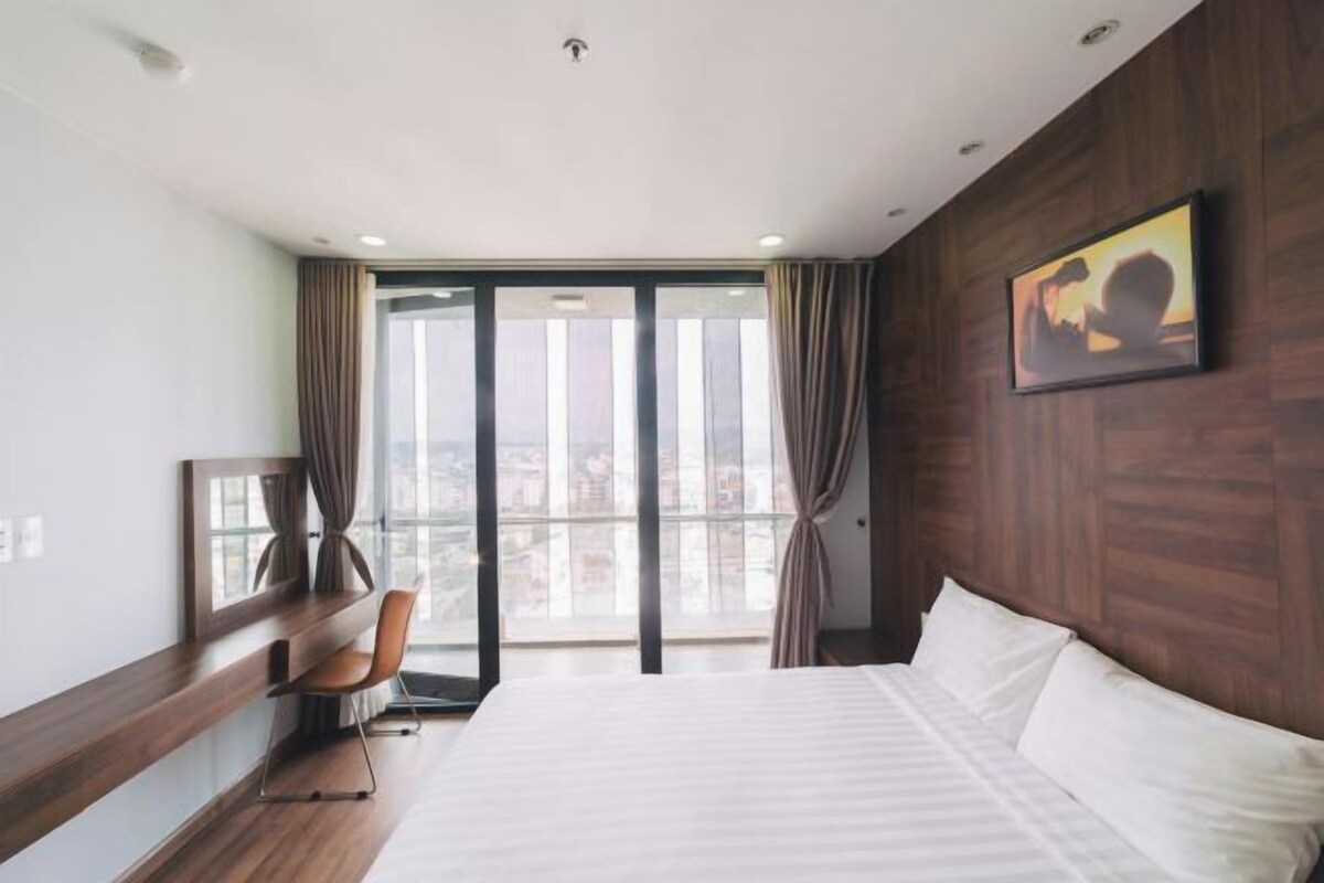 A hotel room with a large bed, a desk, and a view of the city through floor-to-ceiling windows.