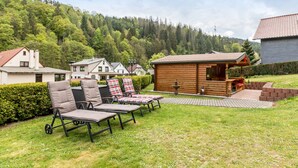 Eine Holzhütte mit Veranda, eine Rasenfläche mit Liegestühlen und ein bewaldeter Hügel im Hintergrund.