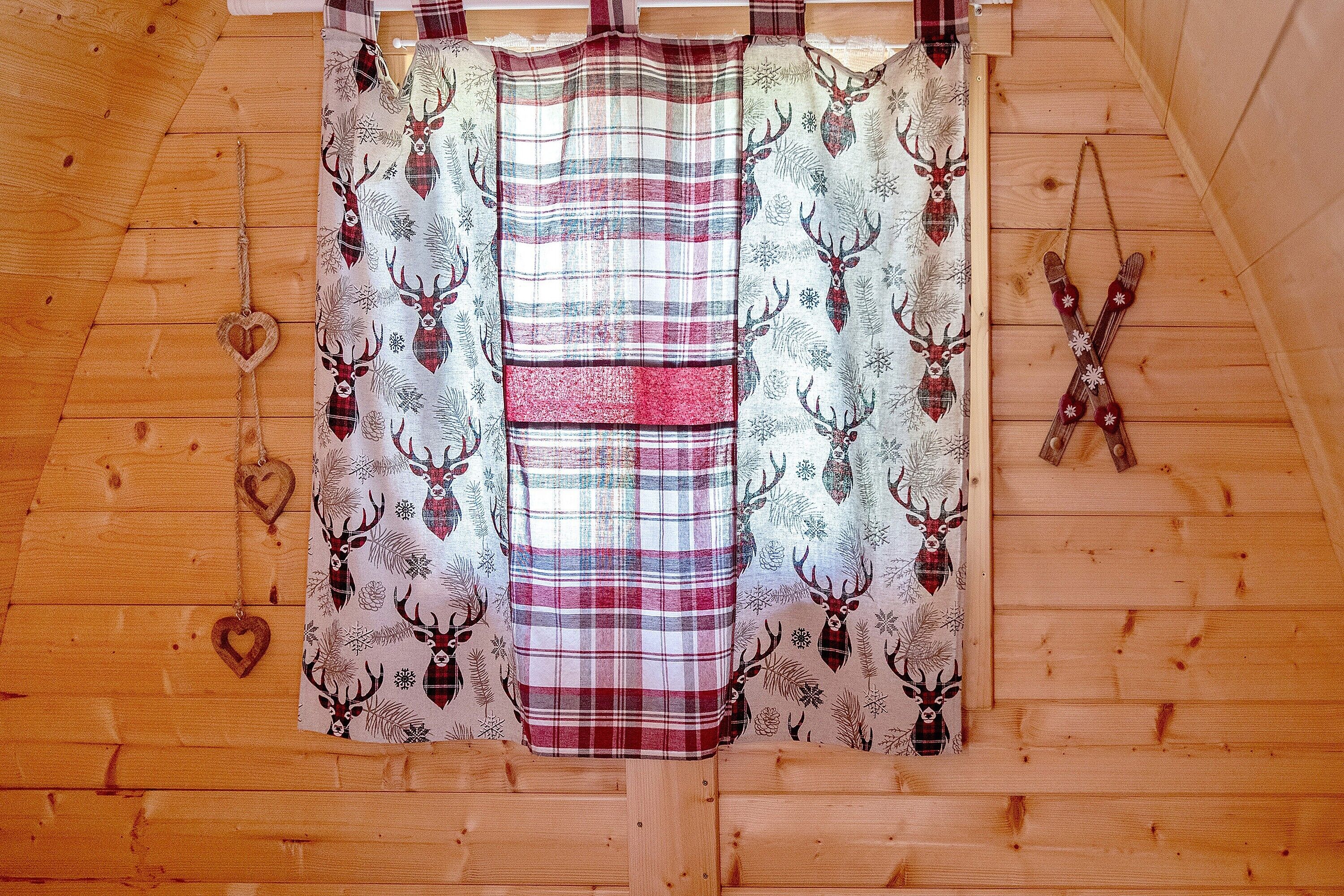 Interior de una cabaña de madera con cortina escocesa que presenta estampados de ciervos y copos de nieve, un adorno con forma de corazón y un par de esquíes cruzados.