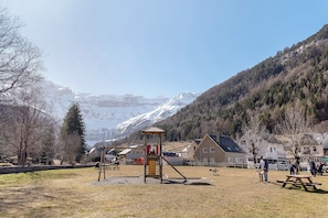 A playground with a slide and climbing structure, surrounded by houses and a mountain backdrop. - Cabane Perce Neige – Gavarnie: Cozy Wooden Cabin in the Heart of the Pyrenees (Gavarnie-Gèdre)