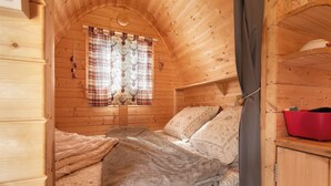 Une pièce chaleureuse, aménagée dans un chalet en bois, avec un lit, une fenêtre avec des rideaux et une étagère sur laquelle est posé un pot rouge.