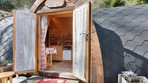 Une structure en bois surmontée d’un toit hexagonal, une porte ouverte donnant sur un espace cuisine et un petit coin salon extérieur.