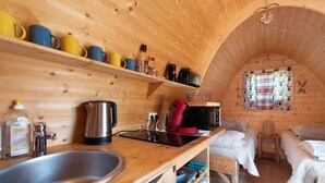 Une cabane en bois chaleureuse, dotée d’un coin cuisine, d’un lit et d’une fenêtre avec des rideaux.
