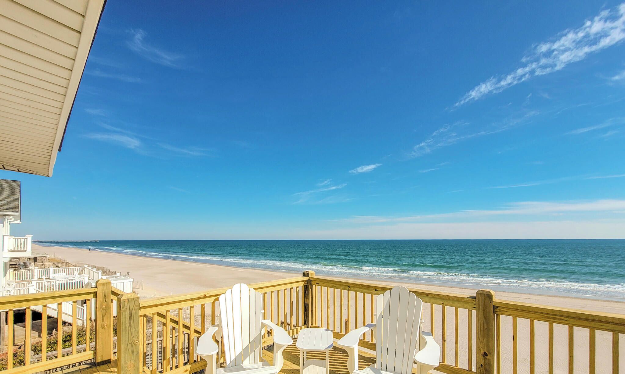 Een houten terras met witte Adirondack-stoelen met uitzicht op een strand met helderblauw water en een zandstrand.