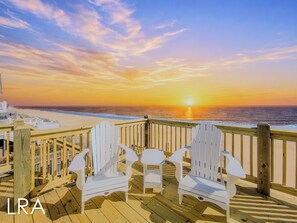 Una terraza de madera con sillas Adirondack blancas con vistas a una playa al atardecer.