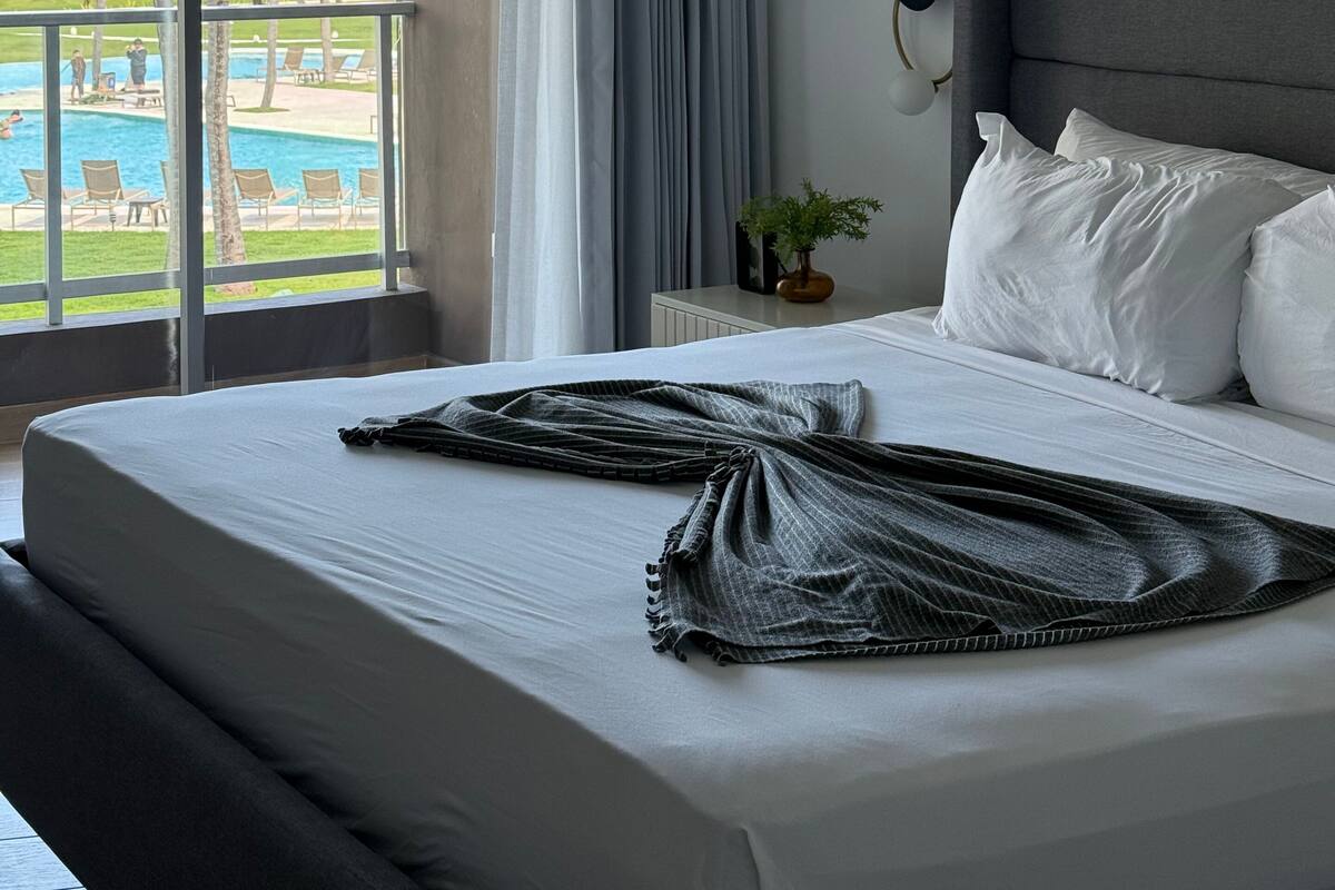 Une chambre d’hôtel avec un lit, une table de chevet, une plante en pot et une vue sur une piscine et des palmiers.