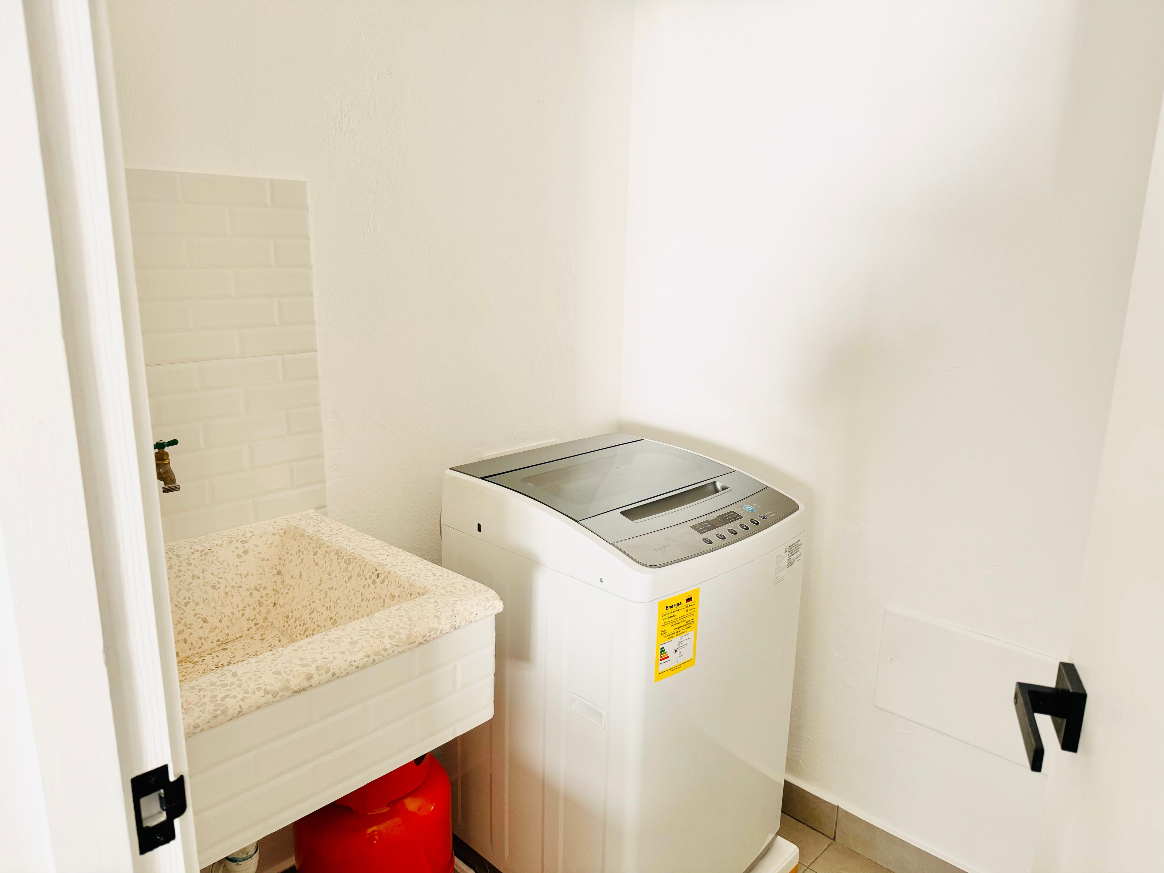 A compact room with a white tiled wall, a small countertop, and a white appliance, possibly a coffee maker or water dispenser, with a red container underneath.
