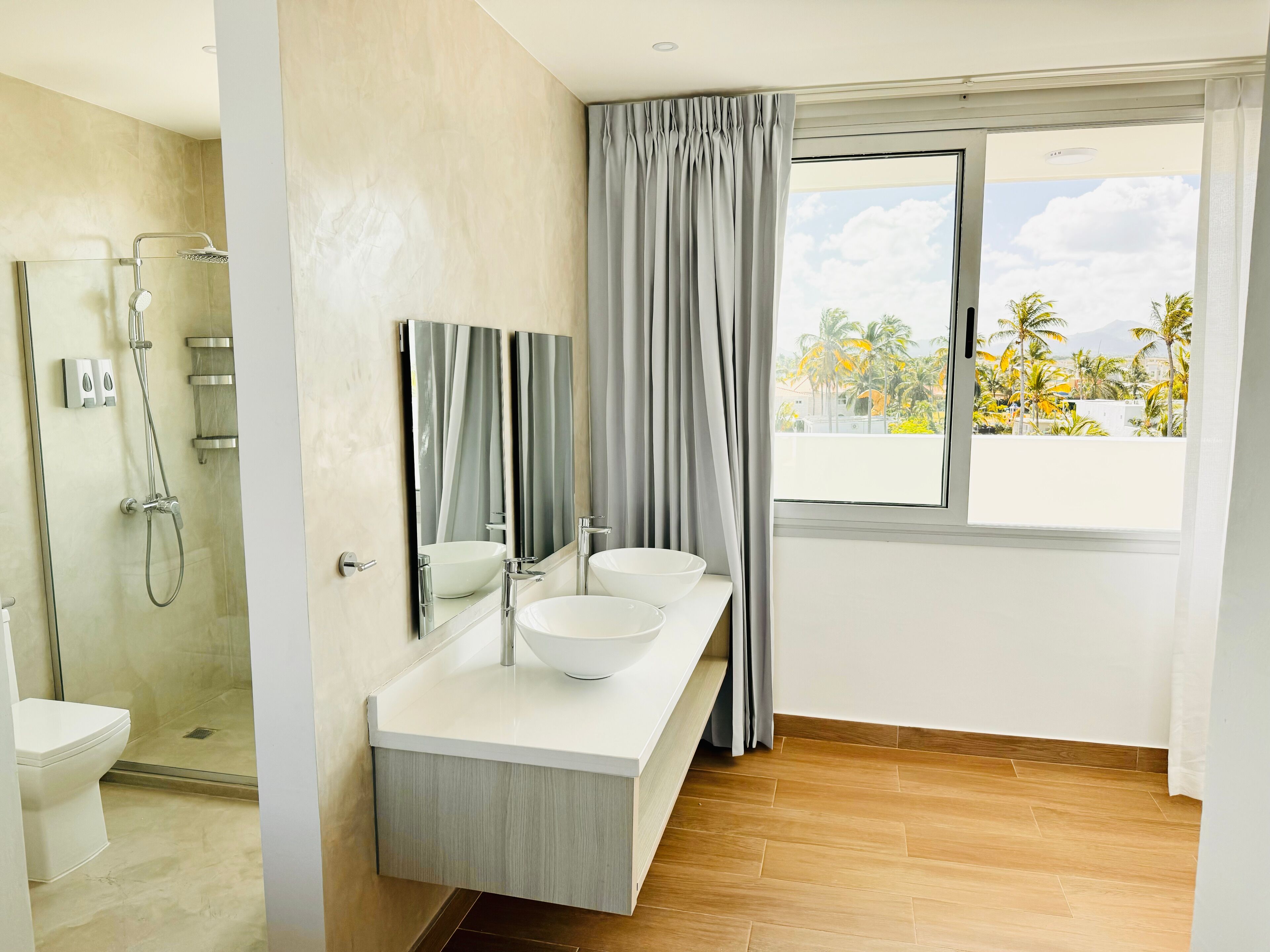 A modern bathroom with a glass-enclosed shower, a large mirror, two sinks, and a view of palm trees through a window.