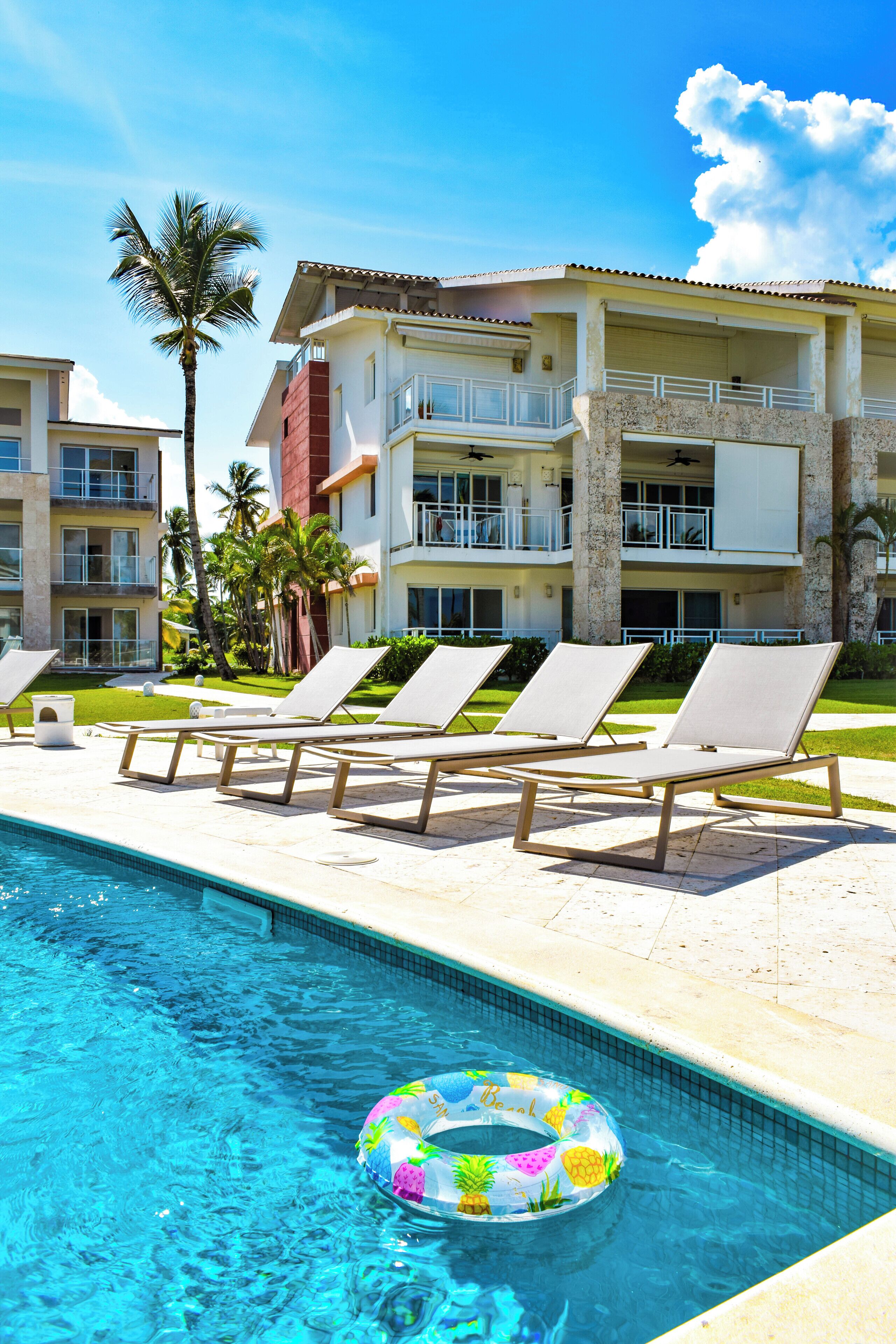 A pool area with lounge chairs and a floating ring, surrounded by a building with balconies and palm trees.