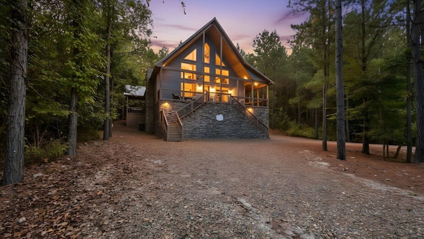 A cabin with a stone foundation, wooden exterior, and large windows. It has a porch with a staircase leading to the entrance.