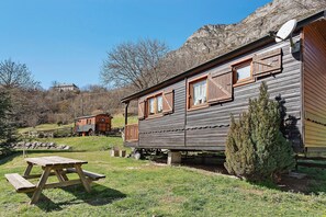 A rustic wooden cabin with a picnic table in front, set in a grassy area with a mountain backdrop. - Roulotte Edelweiss – Unique Wooden Caravan Stay in Gavarnie (Gavarnie-Gèdre)