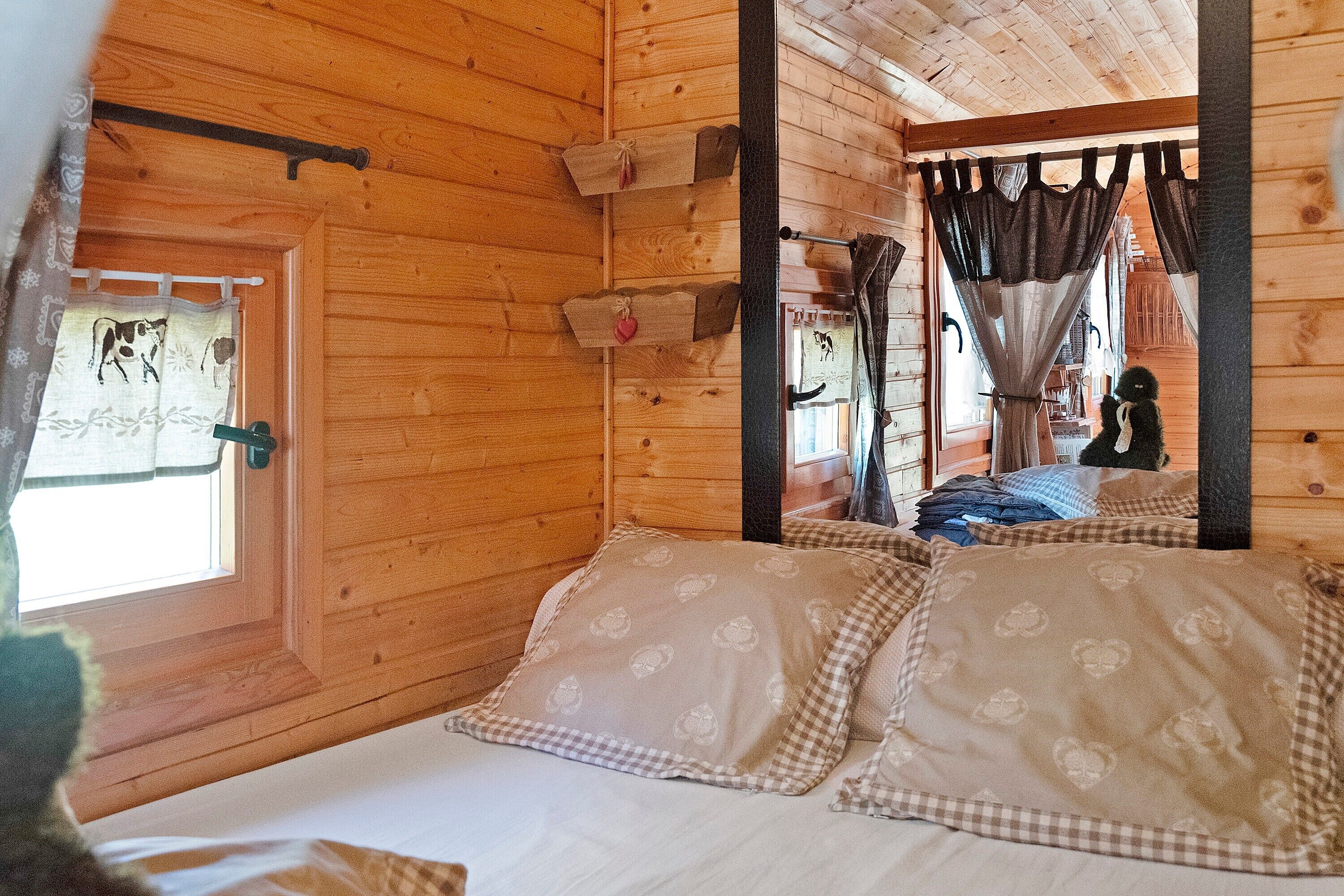 Una habitación de cabaña de madera con una cama, una ventana con cortinas y un pequeño estante con un adorno con forma de corazón.