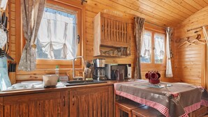 Interior de una cabaña de madera con zona de cocina, armarios de madera, una mesa con mantel a cuadros y una ventana con cortinas.