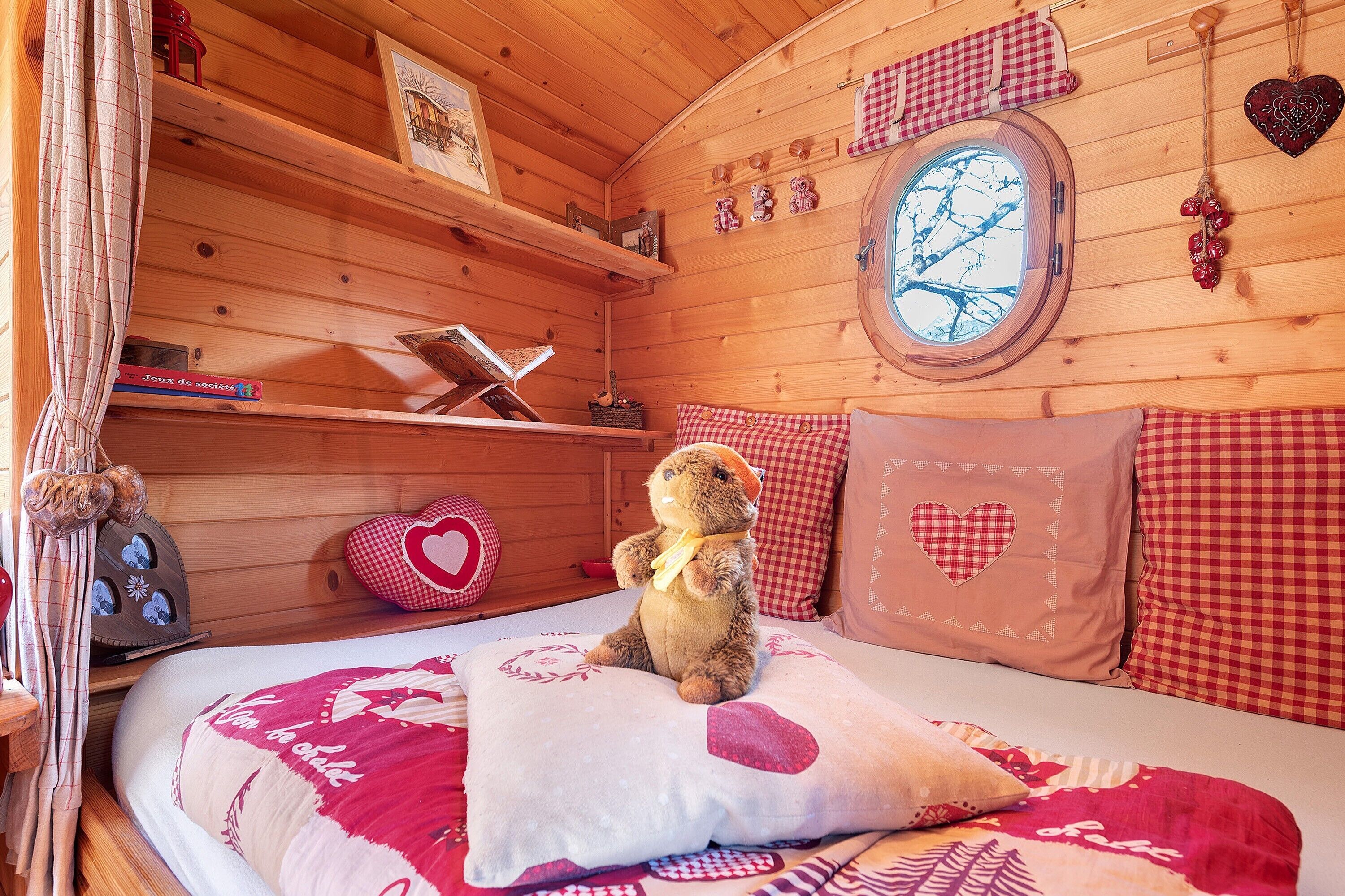 Acogedora habitación tipo cabaña de madera con cama, un oso de peluche, almohadas con forma de corazón y ventana con vistas a árboles.
