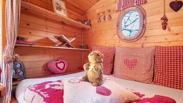 Acogedora habitación tipo cabaña de madera con cama, un oso de peluche, almohadas con forma de corazón y ventana con vistas a árboles.