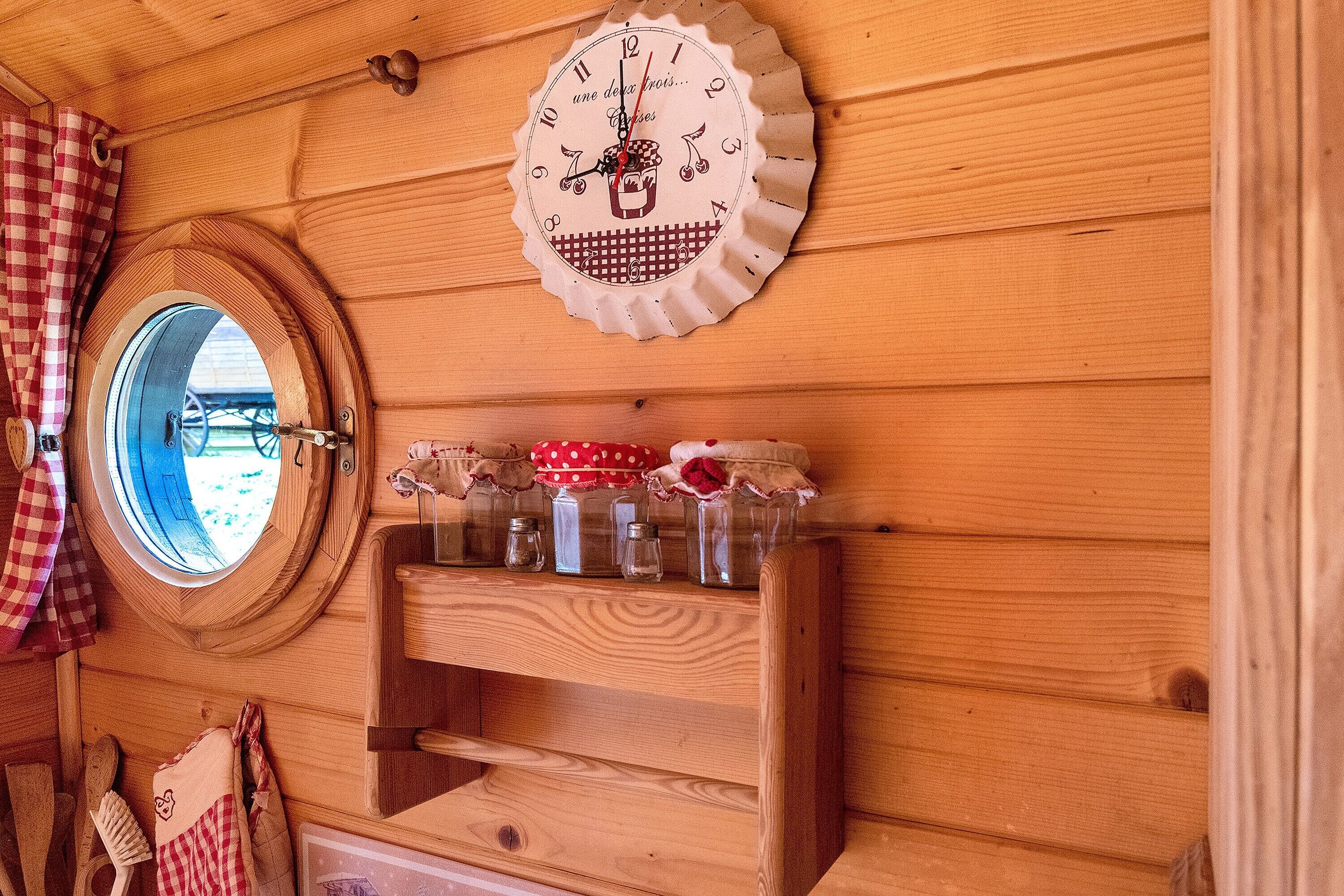 Interior de una cabaña de madera con una ventana redonda, un reloj de pared y una repisa con frascos.