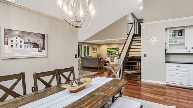 A dining area with a wooden table, chairs, and a chandelier. A staircase leads to a kitchen area with white cabinets. - Sugar Hollow in Chittenden (Rutland)