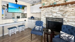 A modern living room with a stone fireplace, a flat-screen TV, and a kitchen area with white cabinets and a wooden countertop. - Admiral s Quarters in Wells (Wells)