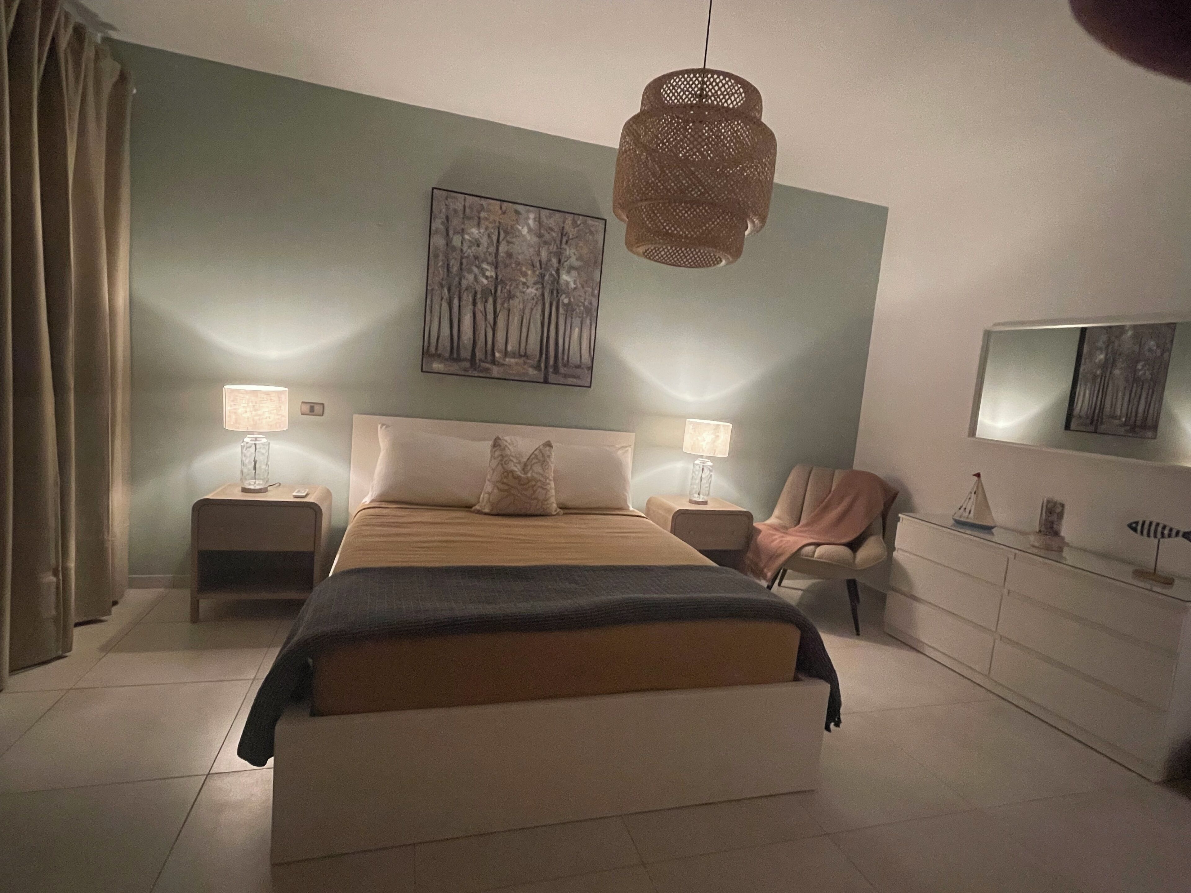 A bedroom with a bed, two bedside tables, a chair, a dresser, and a hanging light fixture.